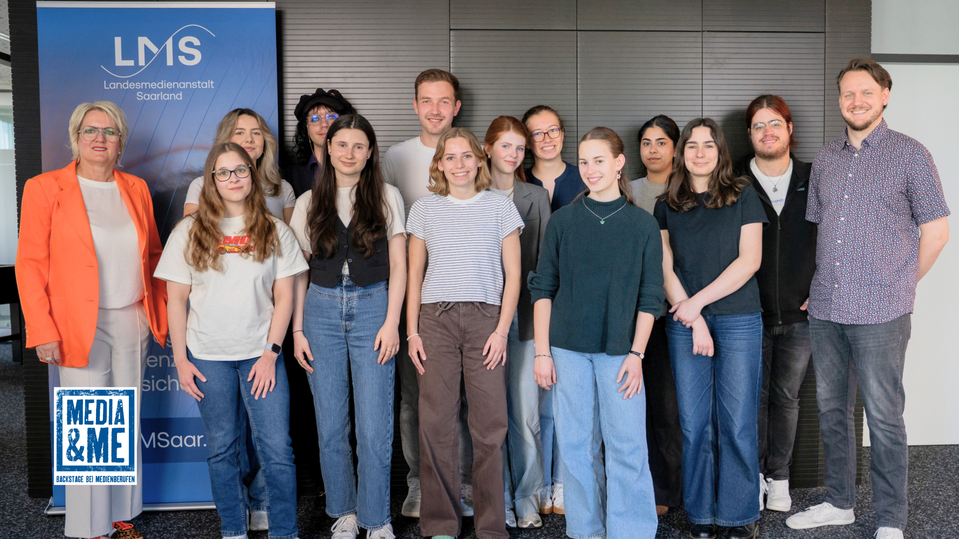 Gruppenfoto der Teilnehmenden und Projektverantwortlichen von Media & Me bei der Landesmedienanstalt Saarland mit LMS-Direktorin Ruth Meyer. Die Gruppe steht vor einem LMS-Banner; unten links ist das Logo von Media & Me eingeblendet.
