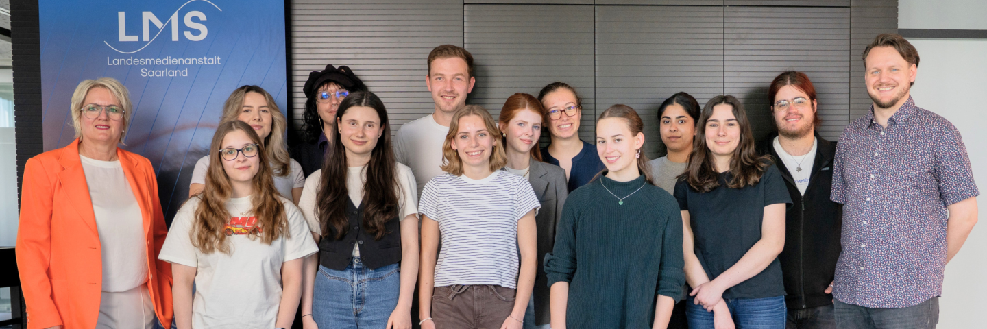 Gruppenfoto der Teilnehmenden und Projektverantwortlichen von Media & Me bei der Landesmedienanstalt Saarland mit LMS-Direktorin Ruth Meyer. Die Gruppe steht vor einem LMS-Banner; unten links ist das Logo von Media & Me eingeblendet.